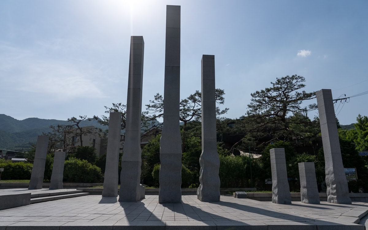 Tall gray stone pillars of the Root of Democracy Sculpture stand on a paved plaza, with trees, mountains, and a bright sky in the background.